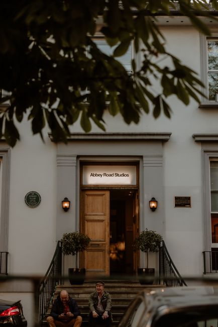 The exterior of a building housing Abbey Road Studios, with a white facade and a set of stone steps leading up to a wooden double door. Above the door is a sign that reads 'Abbey Road Studios,' illuminated by two wall-mounted lamps on either side. Two potted plants are positioned on the landing, one on each side of the entrance. In the foreground, partially obscured by tree branches, are a black car and a portion of a red vehicle parked on the street below. Two individuals are seated on the steps, one wearing a dark jacket and the other in a patterned jacket, possibly involved in a home relocation or a furniture transport process associated with house removals in St John's Wood, as part of professional moving services provided by St John's Wood Removals.