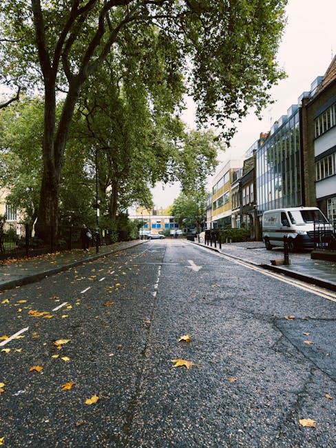A quiet residential street with a wet asphalt road scattered with fallen yellow leaves, flanked by modern multi-storey buildings with large windows on the right and a black metal fence on the left. Tall green trees with dense foliage overhang the street, creating a canopy. Several black bollards line the pavement, and a white van is parked on the right side. In the distance, a delivery truck can be seen on the road, suggesting a possible home relocation or furniture transport activity. The scene is captured during daytime with overcast weather, indicating an environment suitable for interior packing and moving preparations, as managed by St John's Wood Removals for house relocation projects such as those described in Abbey Road studio move tips.